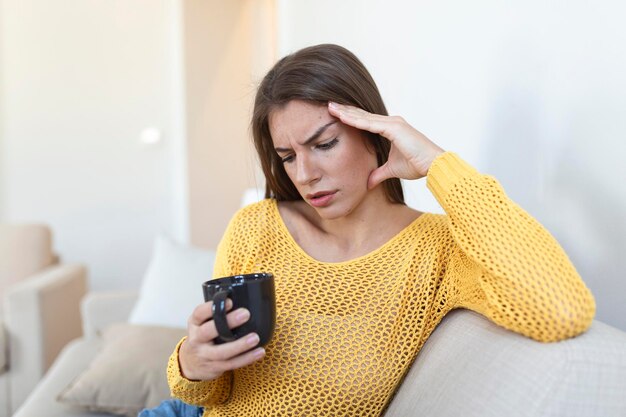 Mujer joven deprimida con dolor de cabeza mientras bebe café en el sofá en casa