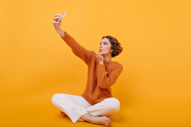 Mujer joven descalza en suéter enviando beso al aire mientras hace selfie