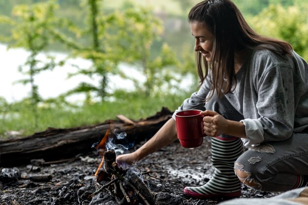 Mujer joven descansando cerca del fuego con una taza de bebida caliente en el bosque cerca del río.