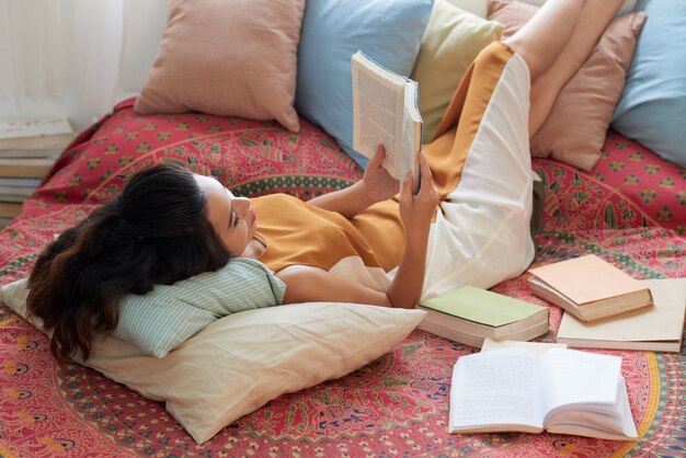 Mujer joven descansando con el libro en la cama con las piernas sobre las almohadas