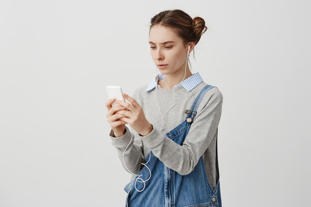 Mujer joven desconcertada en auriculares frunciendo el ceño teniendo problemas con su dispositivo. La encantadora productora está decepcionada por la mala conexión a Internet en su teléfono. Concepto de tecnología