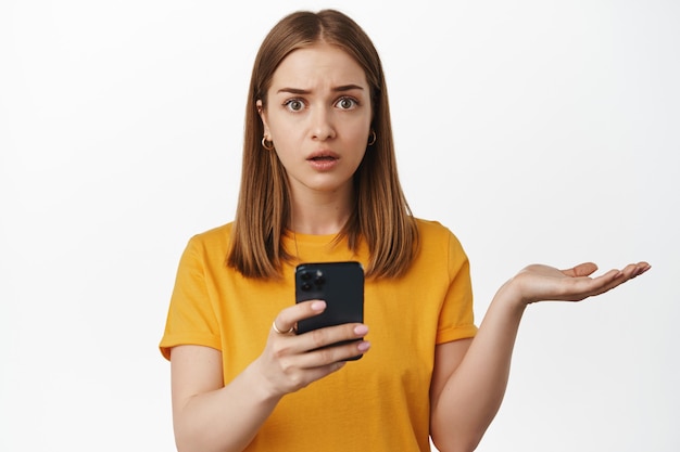 Mujer joven desconcertada sosteniendo el teléfono, no puedo entender, encogiéndose de hombros y luciendo despistada preocupada después de usar la aplicación de teléfono inteligente, de pie sobre una pared blanca