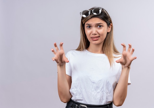 Foto gratuita una mujer joven descontenta en camiseta blanca con gafas de sol mostrando hacer un gesto de garra como un gato mientras mira en una pared blanca