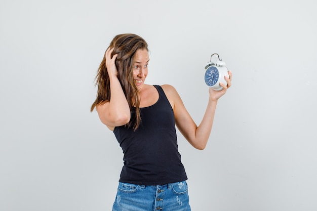 Foto gratuita mujer joven con despertador en camiseta, pantalones cortos y mirando ansioso. vista frontal.