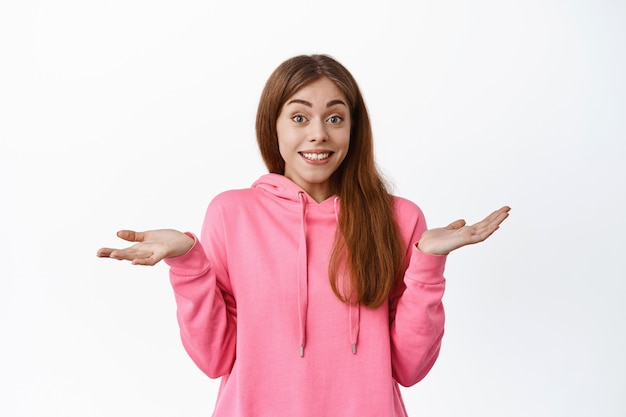 Mujer joven despistada encogiéndose de hombros y sonriendo, no puedo decir, no puedo entender algo, de pie perplejo sobre la pared blanca