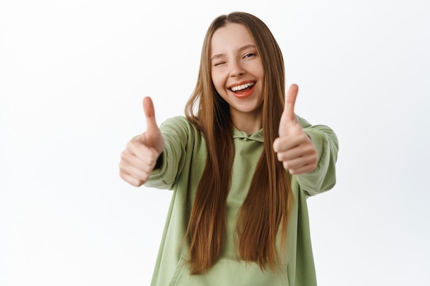 Mujer joven despreocupada estirar los pulgares hacia arriba y guiñarte un ojo, elogiar el buen trabajo, el excelente trabajo, recomendar algo genial, dar me gusta y decir que sí, de pie sobre una pared blanca