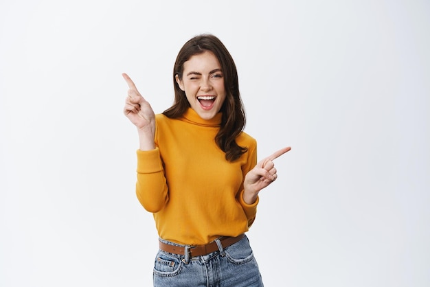 Mujer joven despreocupada guiñando un ojo y sonriendo apuntando de lado a dos ofertas promocionales que muestran publicidad de pie en ropa informal contra fondo blanco