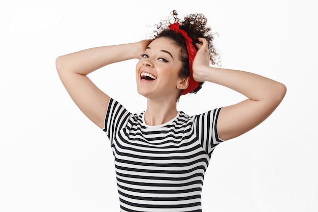 Mujer joven despreocupada con el pelo peinado y la diadema, cogida de la mano en la cabeza y sonriendo, riendo relajada, sintiéndose libre y feliz, de pie contra un fondo blanco.