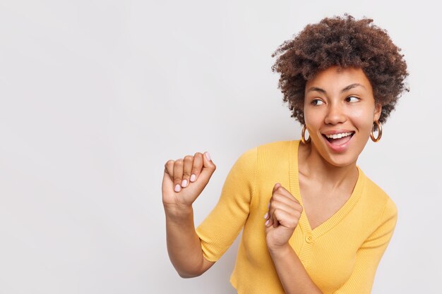 Mujer joven despreocupada positiva sacude los puños se mueve con el ritmo de la música se siente feliz baila y mira hacia otro lado usa un jersey amarillo casual aislado sobre una pared blanca