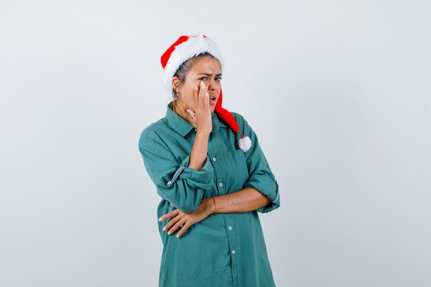 Mujer joven diciendo secreto manteniendo la mano cerca de la boca en camisa, gorro de Papá Noel y mirando con cuidado, vista frontal.