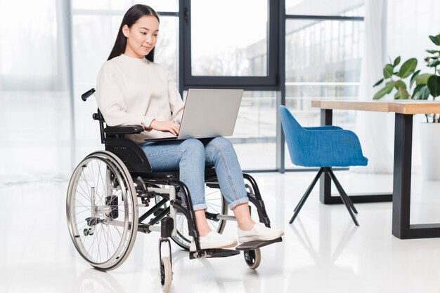 Mujer joven discapacitada sonriente que se sienta en la silla de ruedas usando el ordenador portátil en oficina