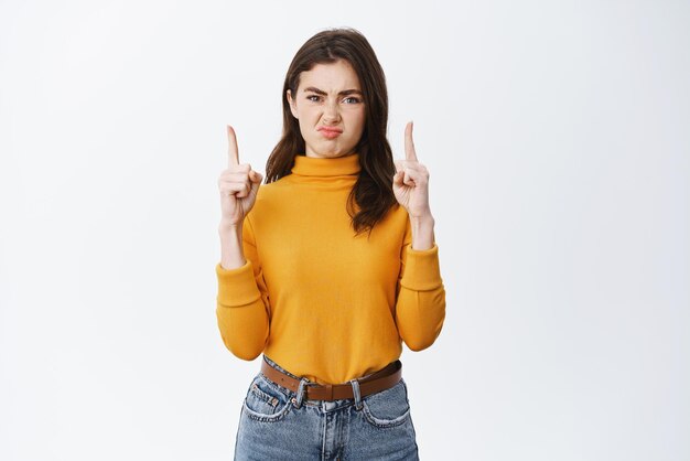 Mujer joven disgustada expresa disgusto señalando con el dedo algo malo haciendo muecas y entrecerrando los ojos decepcionado de pie contra el fondo blanco