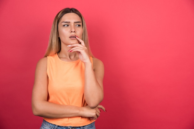 Mujer joven disgustada manteniendo su mano en la barbilla en la pared roja.