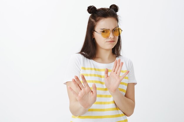 Mujer joven disgustada posando con gafas de sol contra la pared blanca