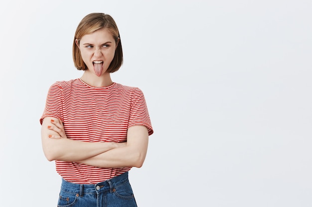 Mujer joven disgustada que hace muecas mostrando la lengua y quejándose por algo malo, mirando a la derecha con mueca molesta