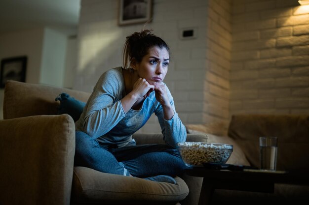 Mujer joven disgustada sentada en un sillón y viendo la televisión a altas horas de la noche en casa