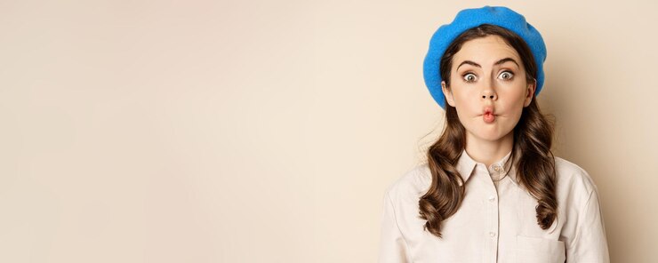 Foto gratuito mujer joven y divertida actuando tonteando con los labios fruncidos y haciendo muecas posando contra un fondo beige con un moderno sombrero azul
