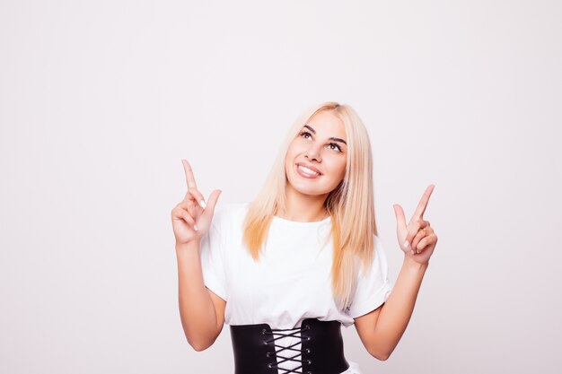 Mujer joven divertida en camisa blanca que señala el dedo hacia arriba aislado. Jovencita rubia haciendo muecas y mirando hacia arriba