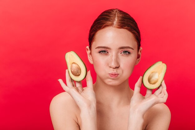 Mujer joven divertida del jengibre que sostiene el aguacate. Vista frontal de una chica guapa aislada sobre fondo rojo.