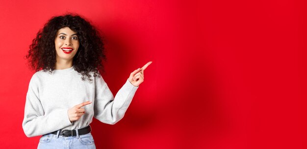 Foto gratuita mujer joven divertida con peinado rizado apuntando con el dedo directamente al logo que parece impresionada y emocionada