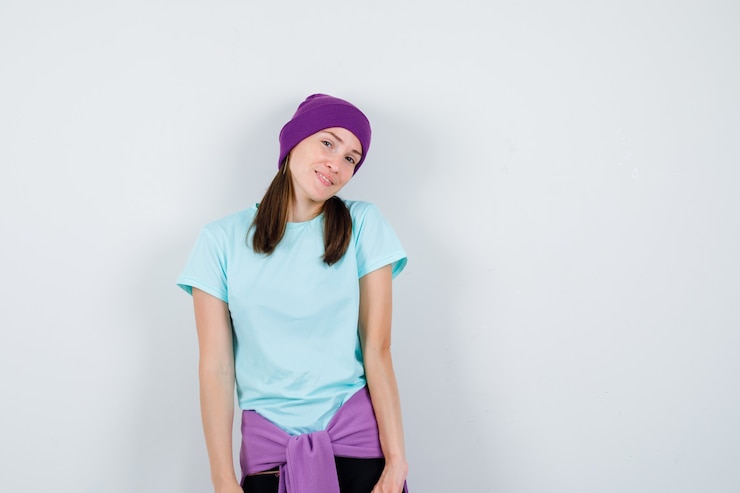 Foto gratuito mujer joven doblando la cabeza mientras posa en camiseta azul, gorro morado y mirando feliz, vista frontal.