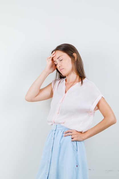 Mujer joven con dolor de cabeza en camiseta, falda y aspecto cansado, vista frontal.