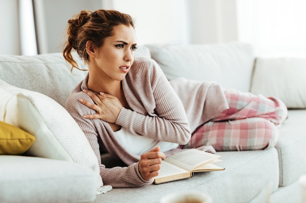Mujer joven con dolor de cuello mientras se relaja en el sofá y lee un libro en casa