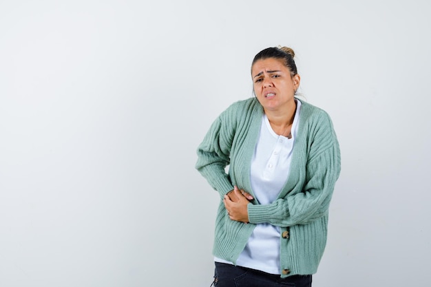 Mujer joven con dolor de estómago, haciendo muecas en camiseta blanca y chaqueta de punto verde menta y mirando molesto