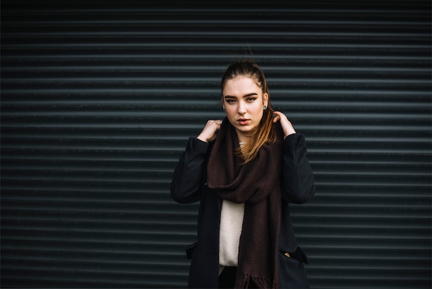 Foto gratuita mujer joven elegante en abrigo con bufanda cerca de la pared de láminas perfiladas