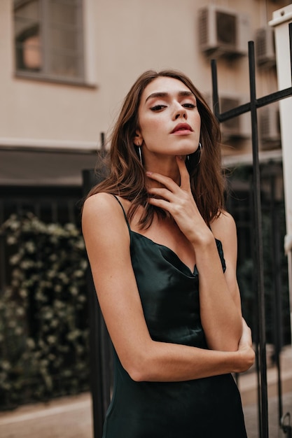 Mujer joven elegante con aretes de maquillaje elegante cabello moreno y vestido de seda verde oscuro posando al aire libre y mirando a la cámara