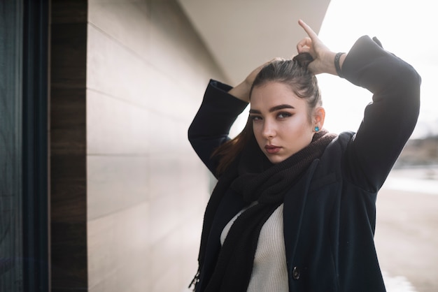 Mujer joven elegante en capa con la bufanda que sostiene el pelo en la calle