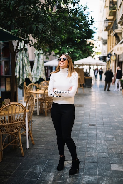 Mujer joven elegante feliz con las gafas de sol cerca del café de la calle en ciudad