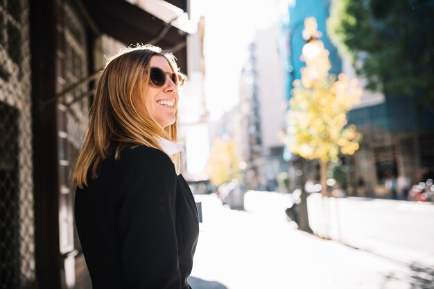 Mujer joven elegante feliz con las gafas de sol en ciudad en día soleado