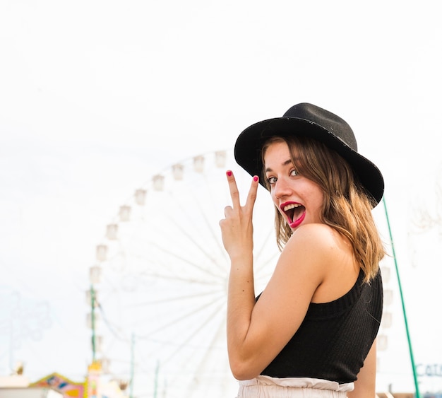Mujer joven elegante feliz que muestra el signo de la paz en el parque de atracciones