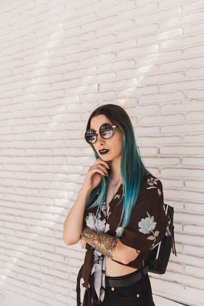 Mujer joven elegante con las gafas de sol que presentan delante de la pared