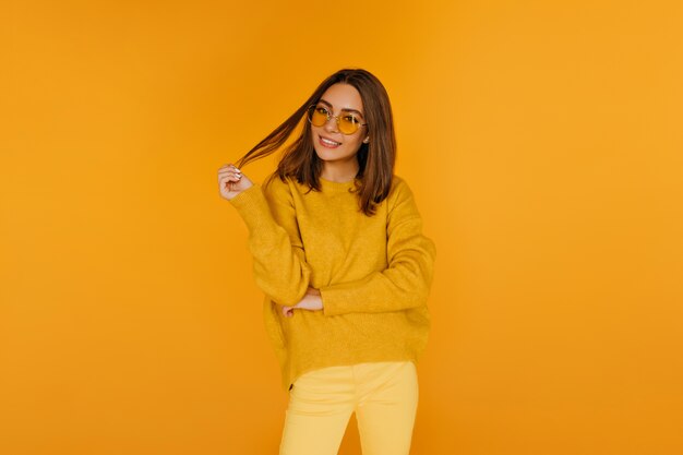 Mujer joven elegante con peinado de moda riendo en la pared amarilla. Filmación en interiores de agraciada niña caucásica en pantalones brillantes.