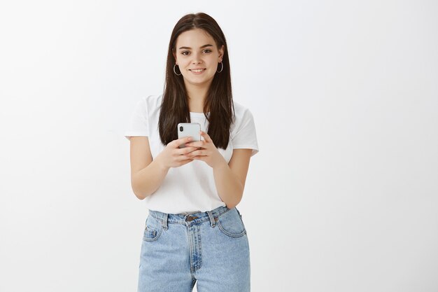 Mujer joven elegante posando con su teléfono contra la pared blanca