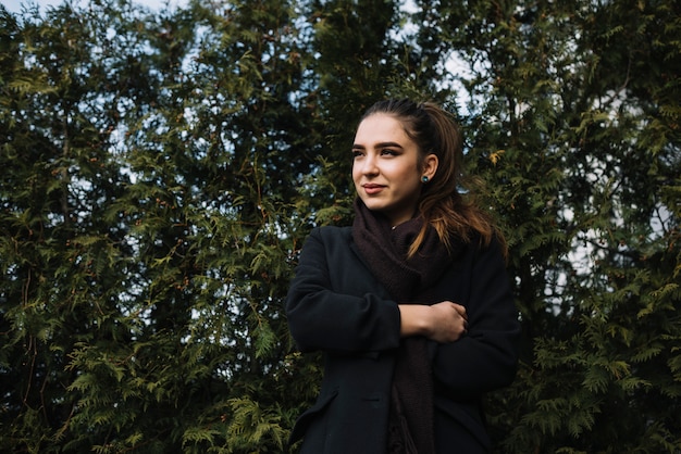 Mujer joven elegante positiva en capa con la bufanda cerca de las plantas coníferas