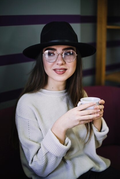 Mujer joven elegante positiva en sombrero y lentes con la taza de bebida
