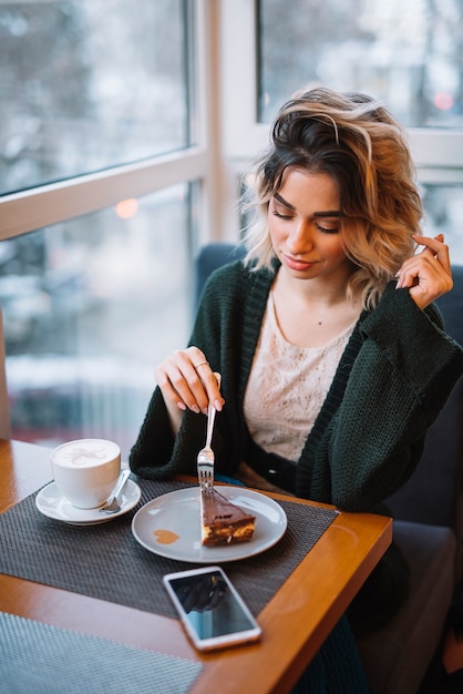 Mujer joven elegante con el postre y la taza de bebida cerca del smartphone en la tabla