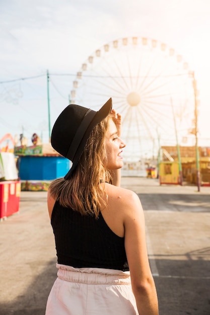 Mujer joven elegante que lleva el sombrero negro en su cabeza que se coloca en el parque de atracciones