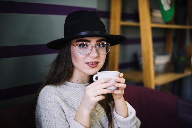 Mujer joven elegante en sombrero y lentes con taza de bebida