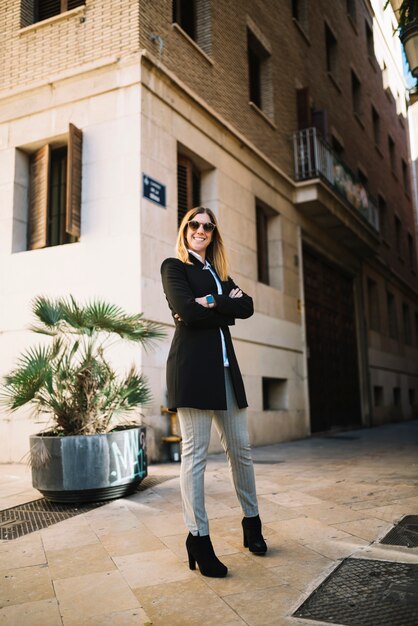 La mujer joven elegante sonriente con las gafas de sol acerca al edificio en la calle