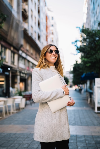 Mujer joven elegante sonriente con las gafas de sol y el ordenador portátil en la calle en ciudad