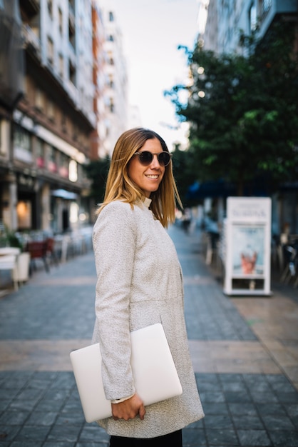 Mujer joven elegante sonriente con las gafas de sol y el ordenador portátil en la calle