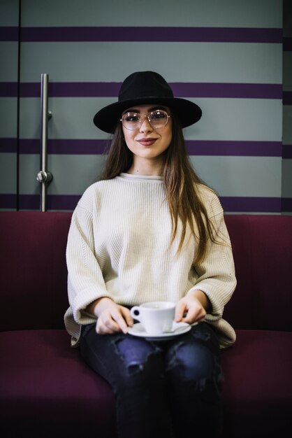 Mujer joven elegante sonriente en sombrero y lentes con la taza de bebida