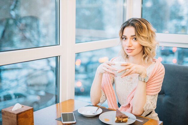 Foto gratuita mujer joven elegante con la taza de bebida cerca del smartphone y el postre en la tabla en café