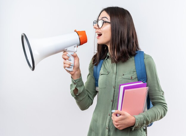 Mujer joven emocionada de la escuela con gafas con mochila sosteniendo libros habla por altavoz