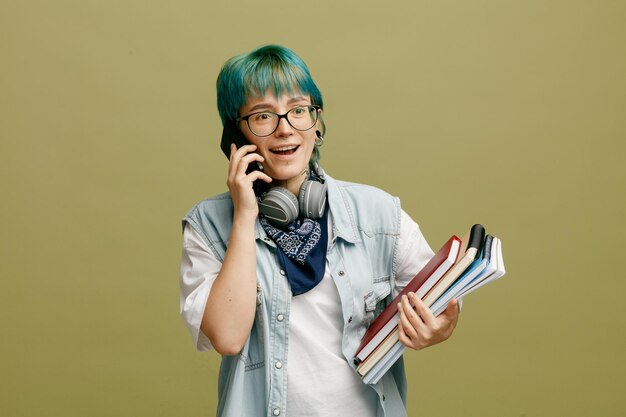 Mujer joven emocionada con gafas, auriculares y pañuelo en el cuello sosteniendo un cuaderno y blocs de notas mirando a un lado mientras habla por teléfono aislado en un fondo verde oliva