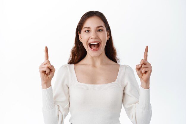 Mujer joven emocionada grita de alegría y hace un anuncio, sonriendo asombrado señalando con el dedo a un lado, mostrando publicidad en la parte superior, de pie feliz contra la pared blanca